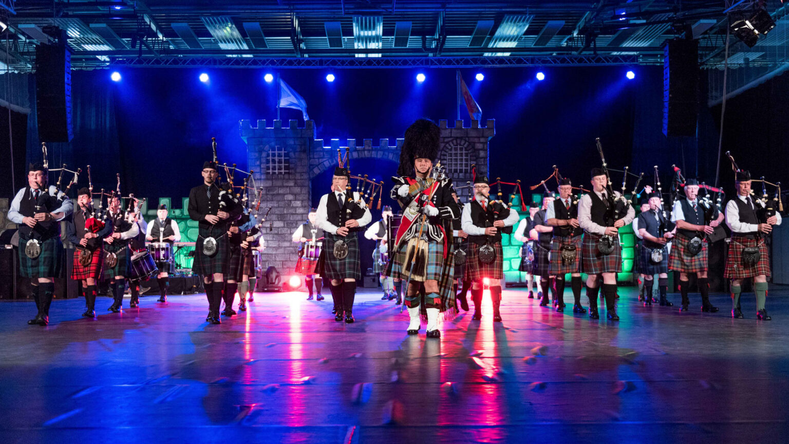 Schottische Dudelsackkapelle mit Pipe Band, Trommlern und Tambourmajor bei einem Konzert auf einer beleuchteten Bühne mit Burgkulisse.